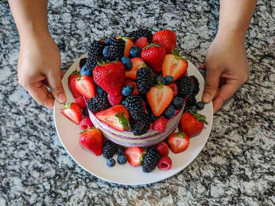 2 hand holding a white circle plate of a cake covered in fresh berries with a grey black marble background