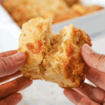 Hand opening a green chile cheddar biscuit and a square pan of the biscuits in the background