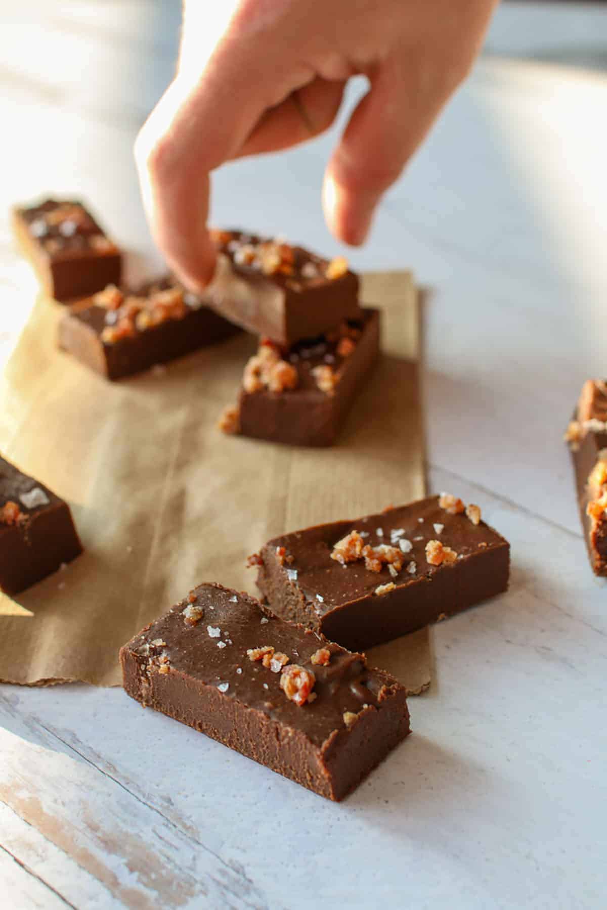 Bacon Fat Chocolate Fudge sliced long with a hand reaching one piece in the back. Fudge is on a piece of paper lunch bag.