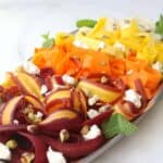 Close Up Angle Rainbow Carrot Ribbon Salad on oval platter with mint on side, topped with feta and pistachios. Carrots in ombree from purple to orange to yellow to white