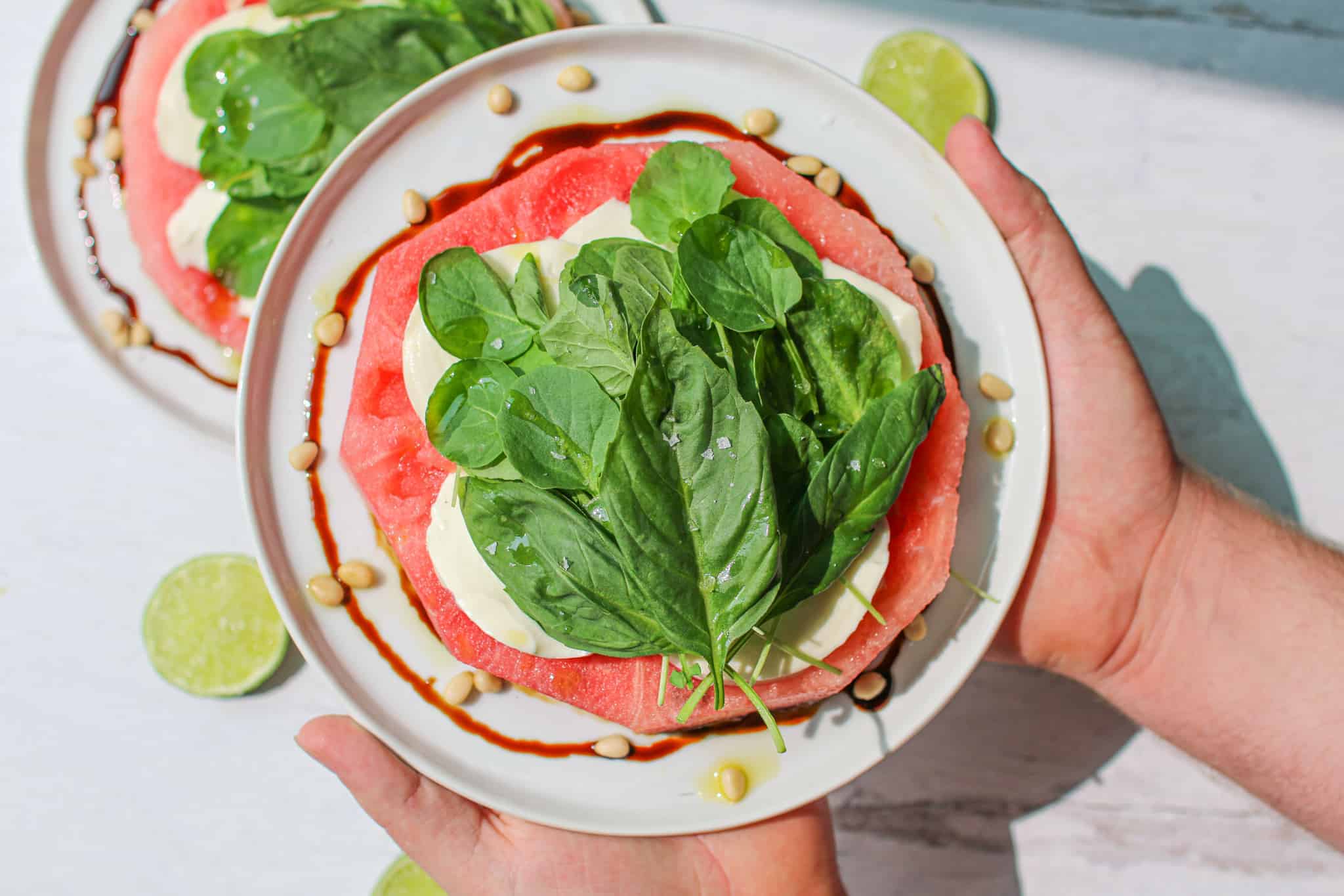 Hands holding up a Fresh Watermelon Mozzarella Salad with basil on a white plate