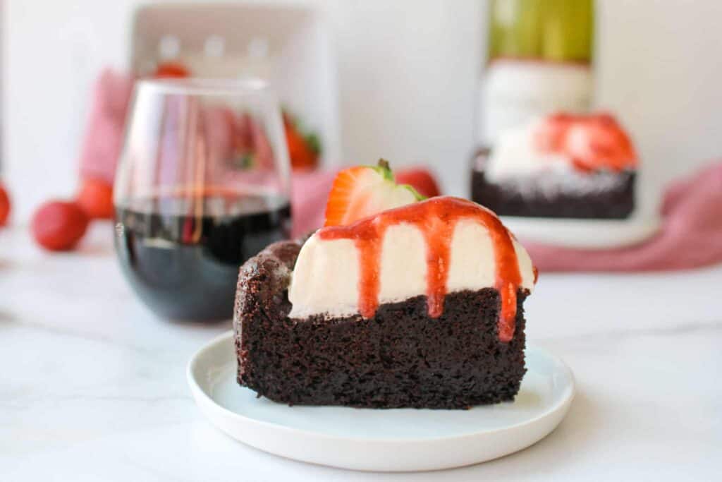 Straight on photo of a slice of leftover red wine chocolate cake with mascarpone whipped cream and strawberry red wine sauce with three drizzles over the side, topped with a strawberry sliced in half. In the background in another slice of cake, red wine bottle, glass half full of red wine, mauve colored cloth and strawberries in a white ceramic basket