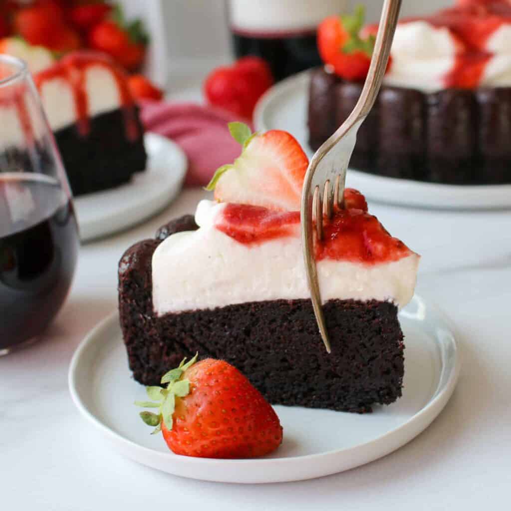 Straight on photo of a slice of leftover red wine chocolate cake topped with mascarpone whipped cream and strawberry red wine sauce with a sliced strawberry and fork taking a bite. The cake is on a small white plate with a whole strawberry. Another slice of cake, the whole cake, a glass of red wine, strawberries and a wine bottle are in the background.