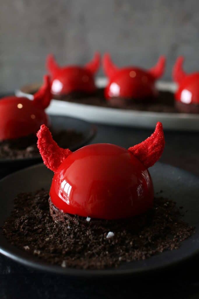 Straight on photo of halloween nutella devil entremet on a black plate, a gray plate has 3 entremets and another entremet on the black plate are in the background. everything sits on a black background and there is a dark grey back