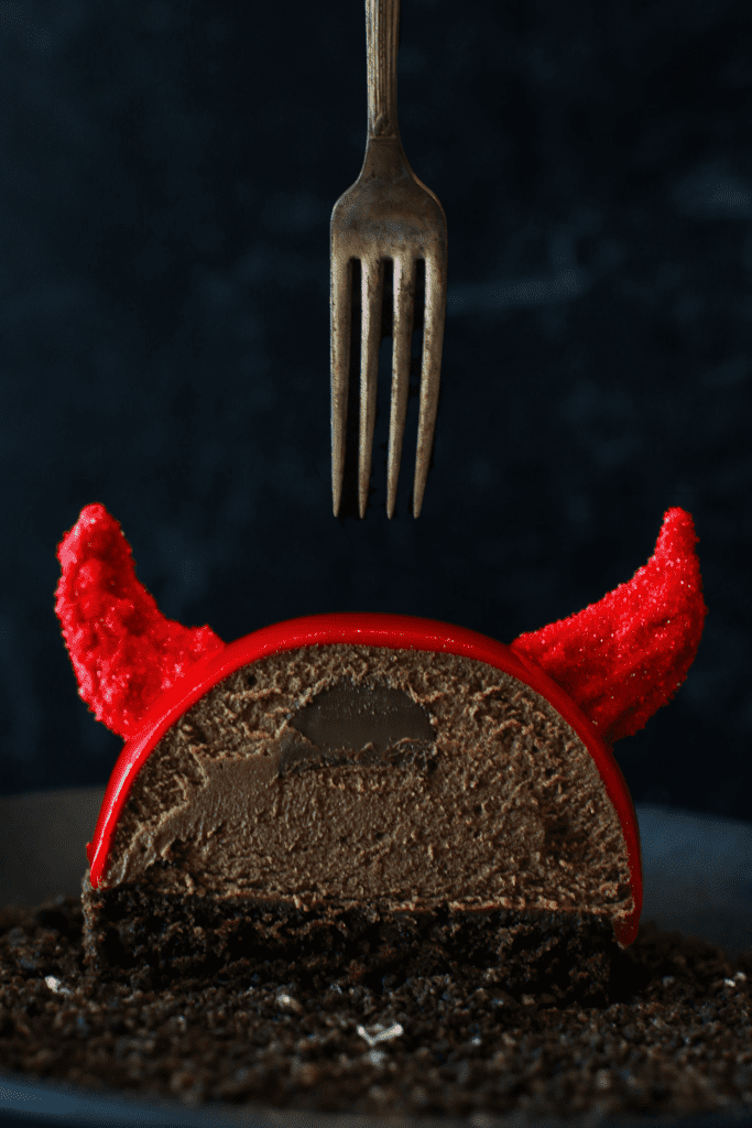 Straight close up on cross-section of a Halloween Nutella Devil Entremet with a fork above. There is a nutella filling surrounded by chocolate nutella mousse semisphere sitting on a cake round of devils food with red glaze and 2 red candy melt horns