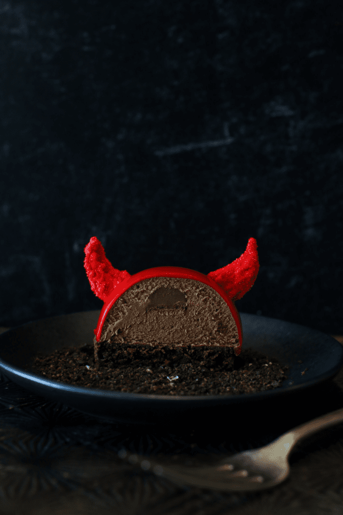 Straight on cross-section of a Halloween Nutella Devil Entremet with a fork in from the plate. There is a nutella filling surrounded by chocolate nutella mousse semisphere sitting on a cake round of devils food with red glaze and 2 red candy melt horns. It sits on a black plate on a dark starburst bottom with a black background