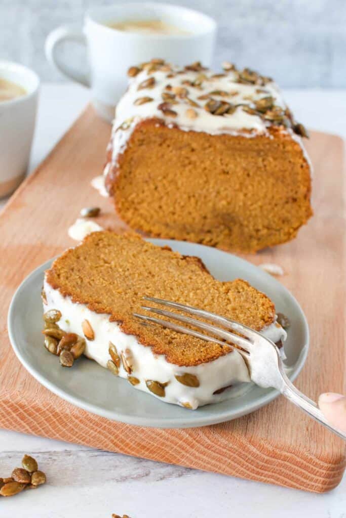 Sliced open pumpkin spice loaf. There's a slice on a small grey plate with a fork going through it. Everything sits on a wooden paddle with a pumpkin spice latte in the background