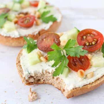 Tzatziki Feta Cream Cheese Spread on two halves of a whole wheat bagel toasted and topped with italian parsley leaves, sliced tomatoes, cucumbers and toasted pine nuts