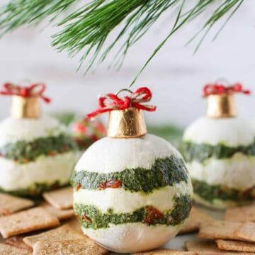 Close up of Ornament Pesto Sun-dried Tomato Cheese Balls on a white place surrounded by wheat thins with a greenery above it in the background