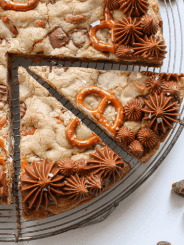 Square photo of close up of Salted Caramel Chocolate Pretzel Cookie Cake with 1/4 of the cookie cake cut into 2 slices on an antique circle cooling rack. with pretzels and baking truffles scattered at the bototm right