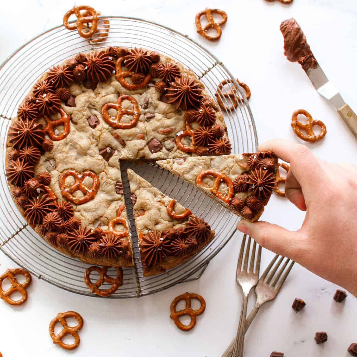 Square photo of Salted Caramel Chocolate Pretzel Cookie Cake with ¼ of the cookie cake cut into 2 slices on an antique circle cooling rack. A hand is picking up 1 of the slices. Mini pretzels scattered around, on the left a small offset spatula with leftover frosting in the top right, bottom right are 2 antique forks crossed slightly and caramel truffle chips scattered in bottom right next to the forks.