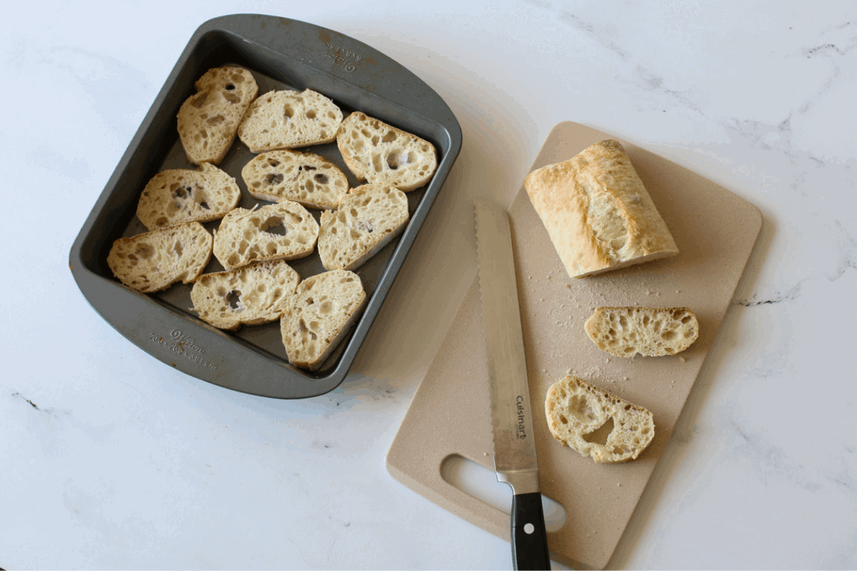 Ciabatta is sliced using a serrated knife. 2 pieces and the end  and the knife are on a small cutting board wit the rest of the sliced bread in an 8x8 baking pan. 