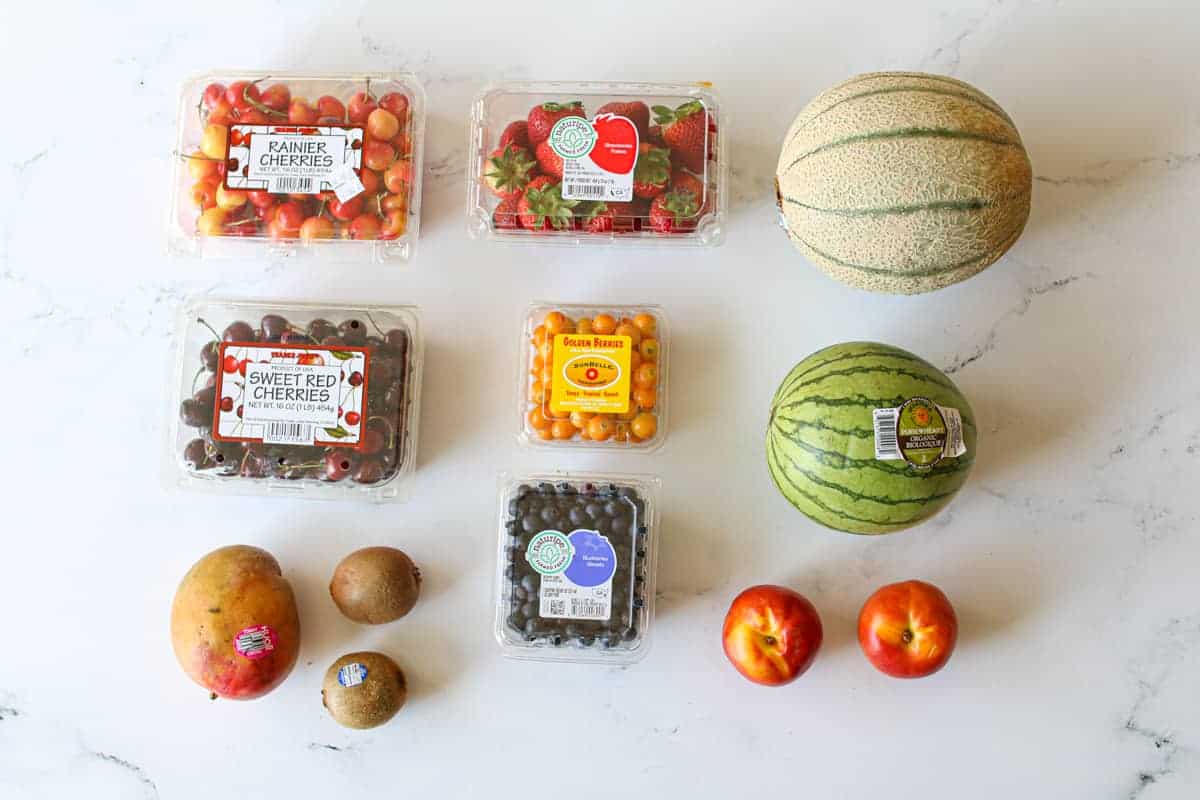 10 variety of fruit on a large white marble background. Rainer cherries, strawberries, goose berries and blueberries are still in their plastic boxes. Starting from top left corner: rainer cherries, strawberries, cantaloupe, next row: sweet red cherries, gooseberries, mini melon, final row with mango, 2 kiwis, blueberries and 2 nectarines 