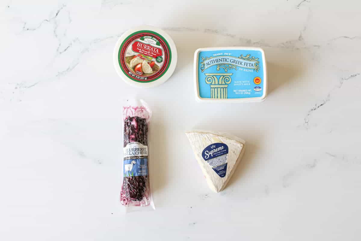 Cheese on white background. Top left is burrata still in its container. Top right is authentic greek feta still in its container. Bottom left is a log of blueberry vanilla goat cheese still its is wrapping. Finally, bottom right is a wedge of supreme  brie still wrapped. 