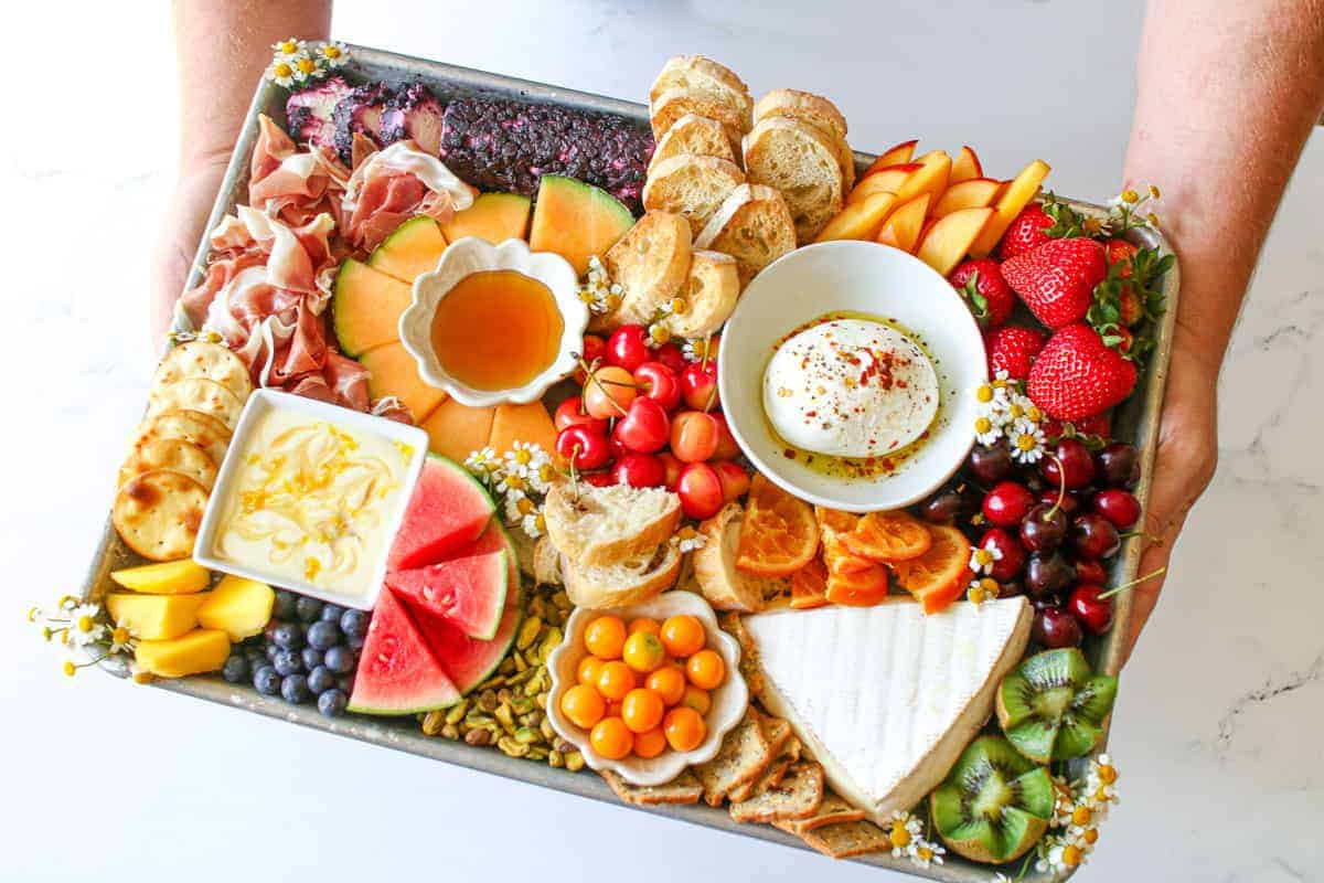 The Trader Joe's Summer Fruit & Cheese Board being held in two hands at an angle on a white marble background.