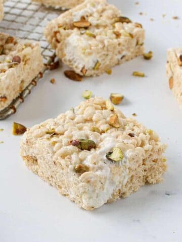 Close up of a whole Salted Pistachio Brown Butter Rice Krispies Treat with multiple Salted Pistachio Brown Butter Rice Krispies Treats in the background and some on an antique potato grater with crushed pistachios surrounding them