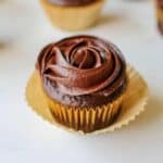 A close up of a chocolate cupcake frosted with sea salt caramel chocolate frosting in a rosette. The cupcake is lined with a brown cupcake liner with another cupcake liner underneath but opened up. The cupcake sits on a white background with more cupcakes in the background