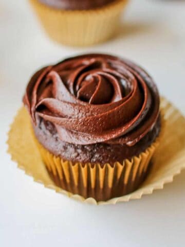 A close up of a chocolate cupcake frosted with sea salt caramel chocolate frosting in a rosette. The cupcake is lined with a brown cupcake liner with another cupcake liner underneath but opened up. The cupcake sits on a white background with more cupcakes in the background