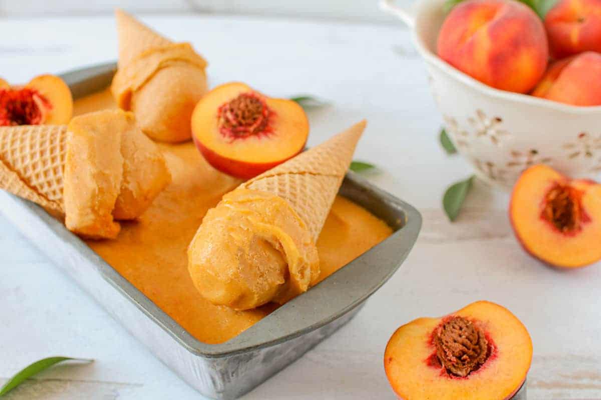 Close up of a scoop of roasted & smoked peach sorbet on a cone sits on top of sorbet in an old loaf pan. Behind the cone are 2 more cones and 2 peach halves. Four whole peaches in a colander to the right. 2 more peach halves are surrounding everything with green leaves.