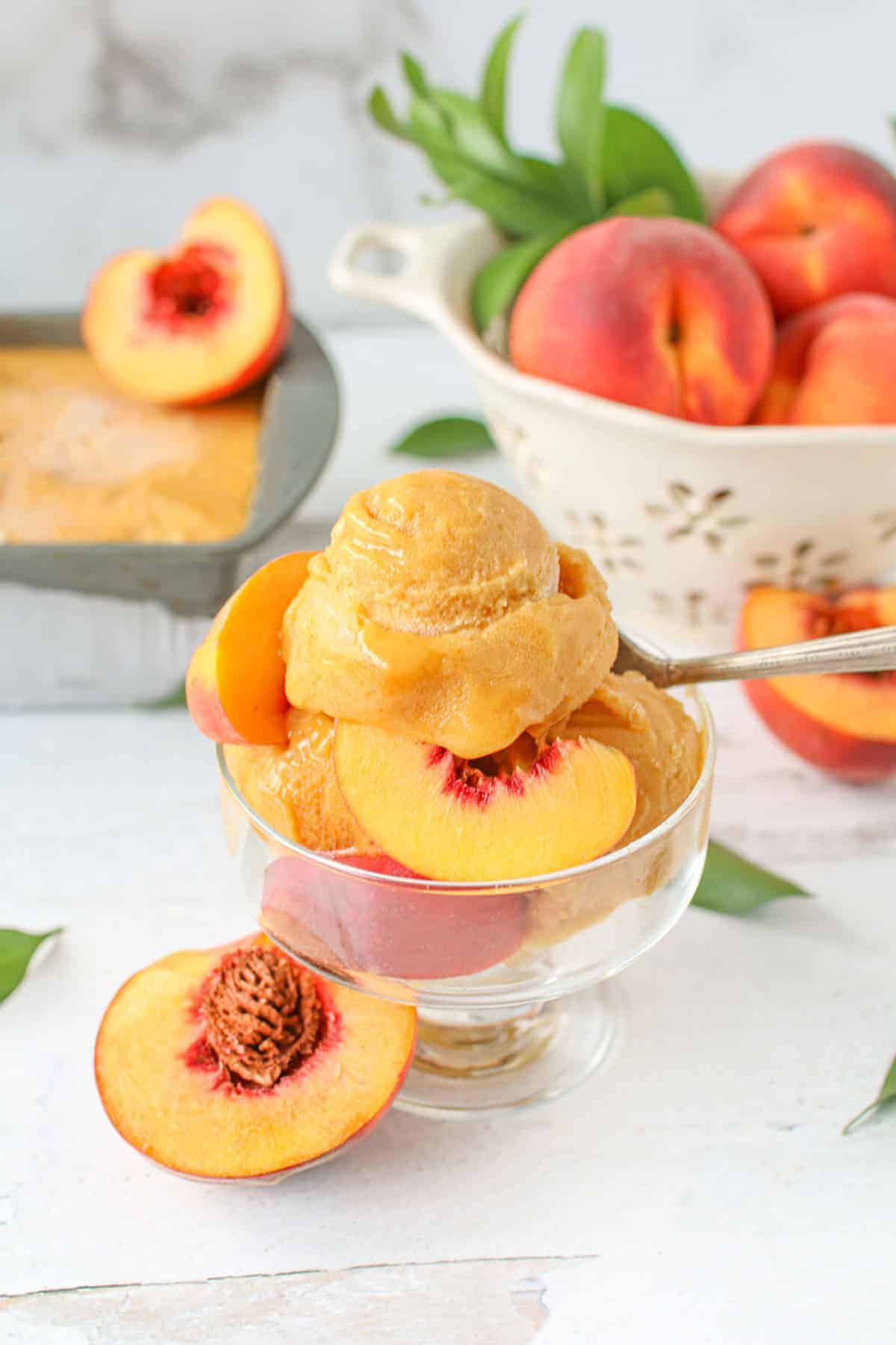 Close up of roasted & smoked peach sorbet in a small glass ice cream bowl with peach slices and a spoon sticking out from the back right of the glass. Behind it on the left is the container of the sorbet in an antique loaf pan with a ½ peach in the top right corner of the container. In the right corner are whole peaches in a cream colored colander with some greenery sticking out.