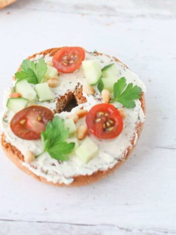 A toasted whole wheat bagel with Tzatziki Feta Cream Cheese Spread topped with sliced cherry tomatoes, diced cucumbers, toasted pine nuts and italian parlsey leaves