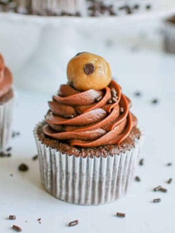 A chocolate cupcake with a chocolate frosting and small cookie dough dall on top with chocolate sprinkles.