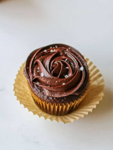 A close up of a chocolate cupcake frosted with sea salt caramel chocolate frosting in a rosette with flaky sea salt on top. The cupcake is lined with a brown cupcake liner with another cupcake liner underneath but opened up. The cupcake sits on a white background with cupcakes cutoff in the background