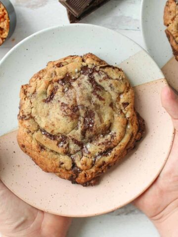 A large cookie is one a white and pink plate with two hands holding it