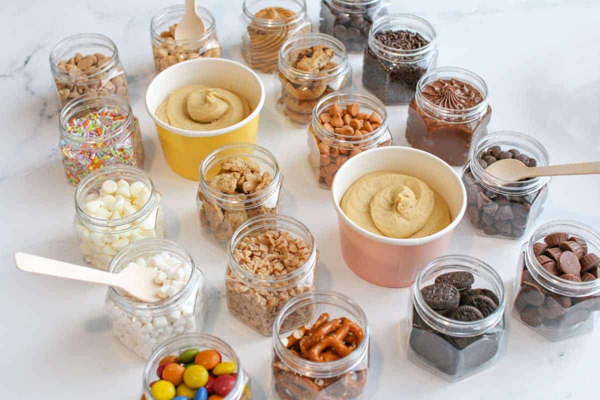 2 small containers with edible cookie dough surrounded by small clear jars with different toppings on a white marble background.