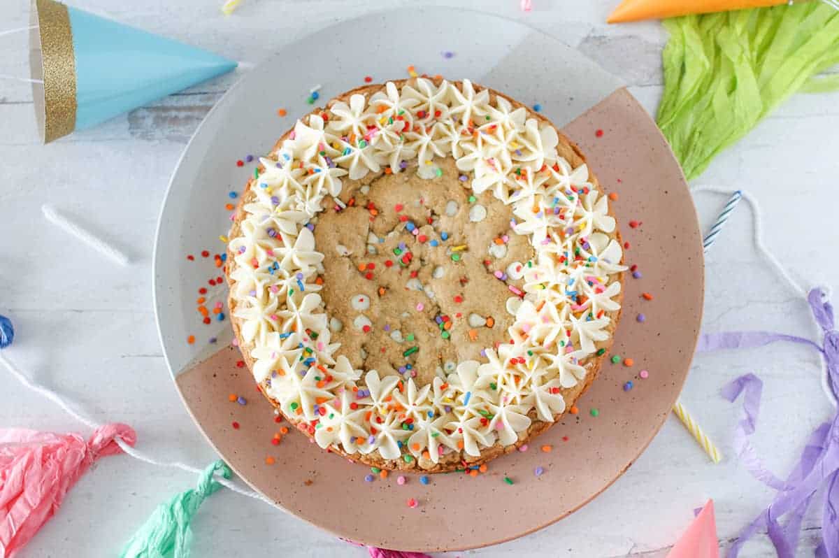 A whole funfetti cookie cake with vanilla frosted border on a pink and white platter with party supplies surrounding the platter.