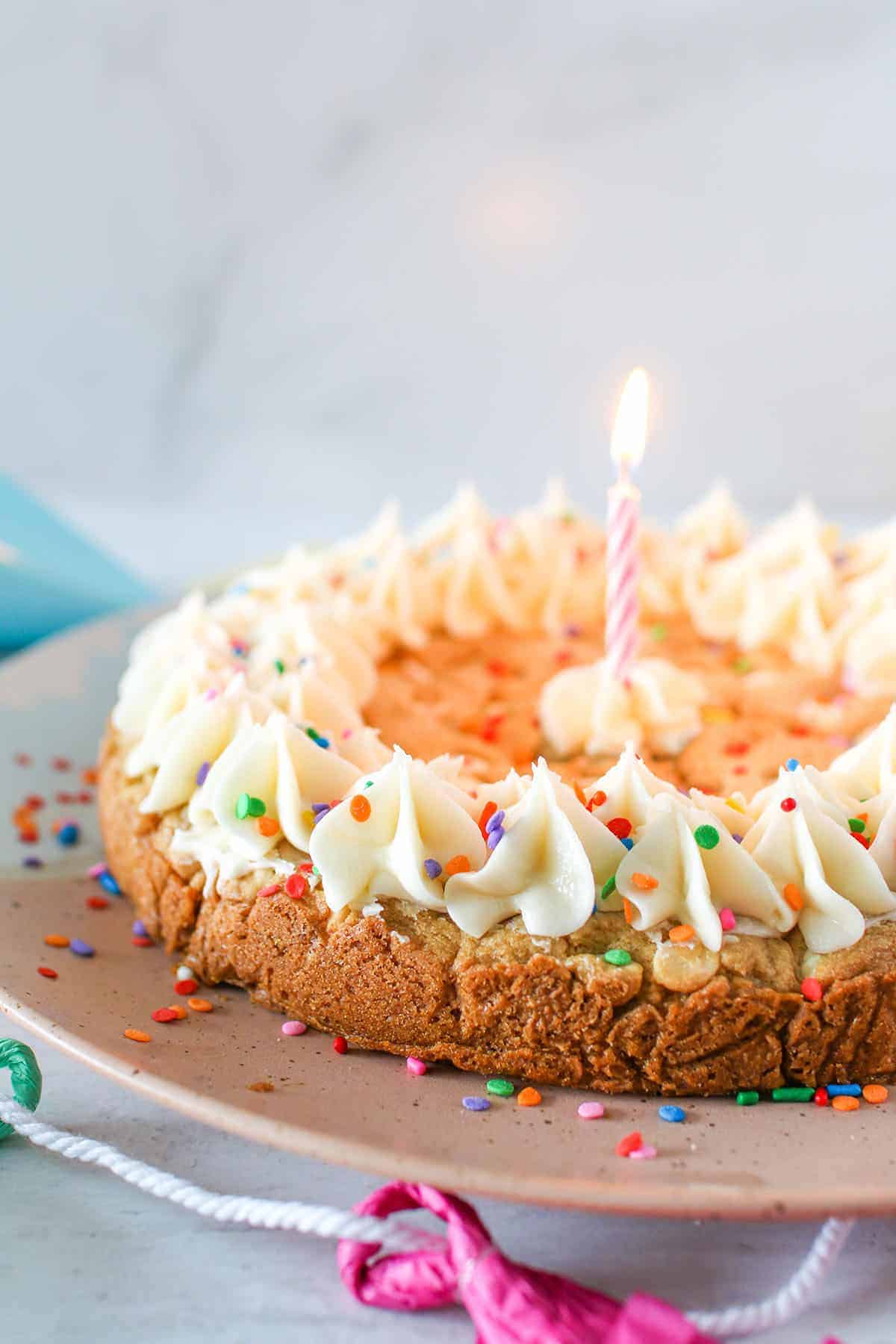 A frosted cookie cake on a white & pink platter with a pink candle lit in the middle. The cookie cake is surrounded by party supplies.