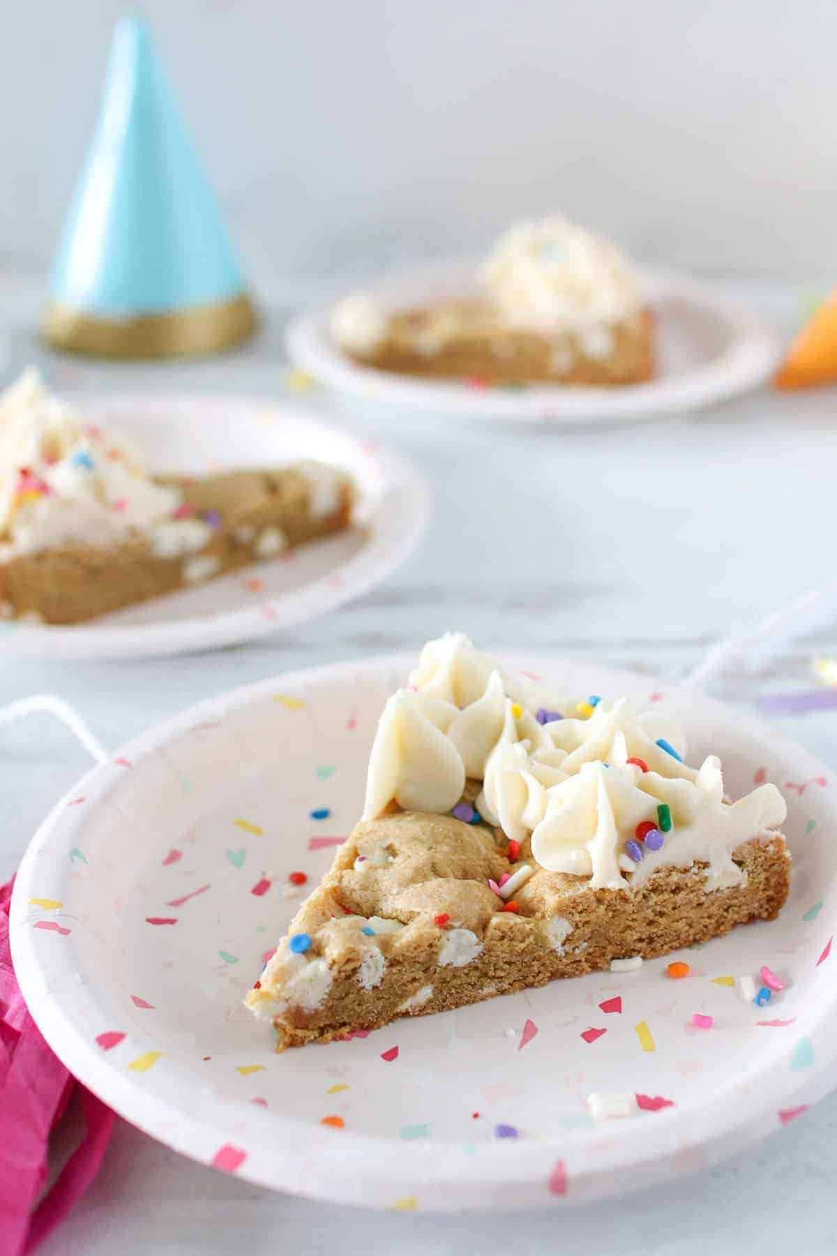 Cookie cake with vanilla frosted border cut into triangles. A small paper plate each has a cookie cake slice. Party supplies are surrounding.