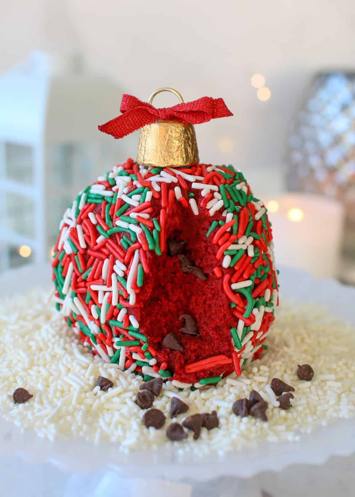 Christmas Ornament Red Velvet Cheese Balls cut into with mini chocolate chips following out, on a bed of white jimmy sprinkles with white, silver and fairy lights in the background