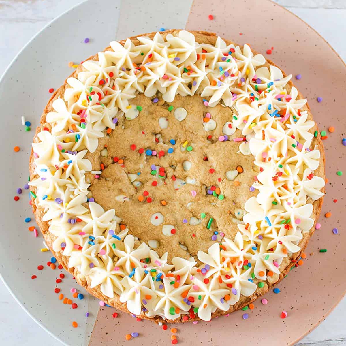 Whole Funfetti Chip Cookie Cake with a vanilla buttercream border and round colorful sprinkles. The cookie cake sits on a white and pink circle platter.