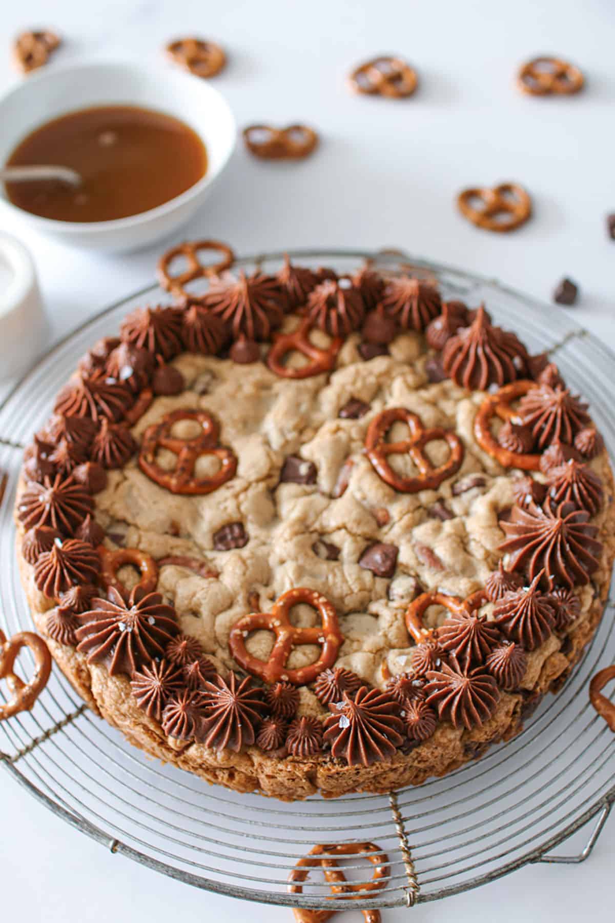 Angled photo of Salted Caramel Chocolate Pretzel Cookie Cake on a large antique silver circle cooling rack with a small bowl of caramel with a spoon in the back left and lots of pretzels scattered around