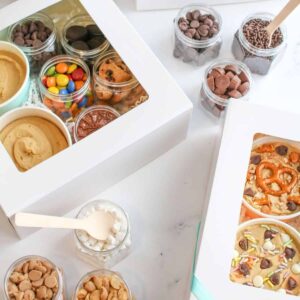 2 boxes with edible cookie dough packs on white marble background. There are small clear jars with different toppoings/mix-ins.