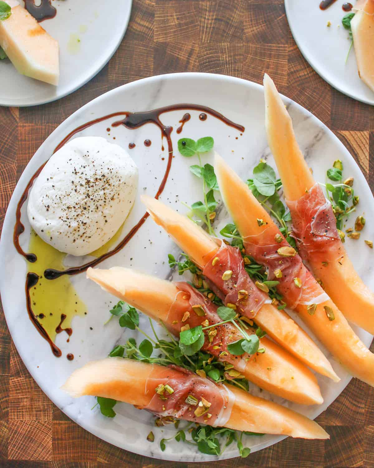 Crenshaw Melon, Burrata and prosciutto on a marble platter with wood cutting brown in the background, two plates off to the side