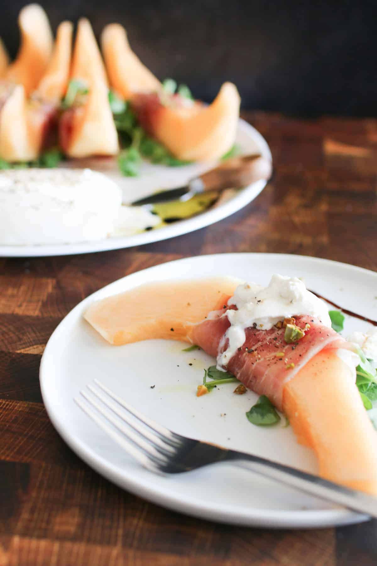 Close up of a slice of Crenshaw Melon, Prosciutto & Burrata on a small white plate with a fork on a butcher block background with the whole platter in the background