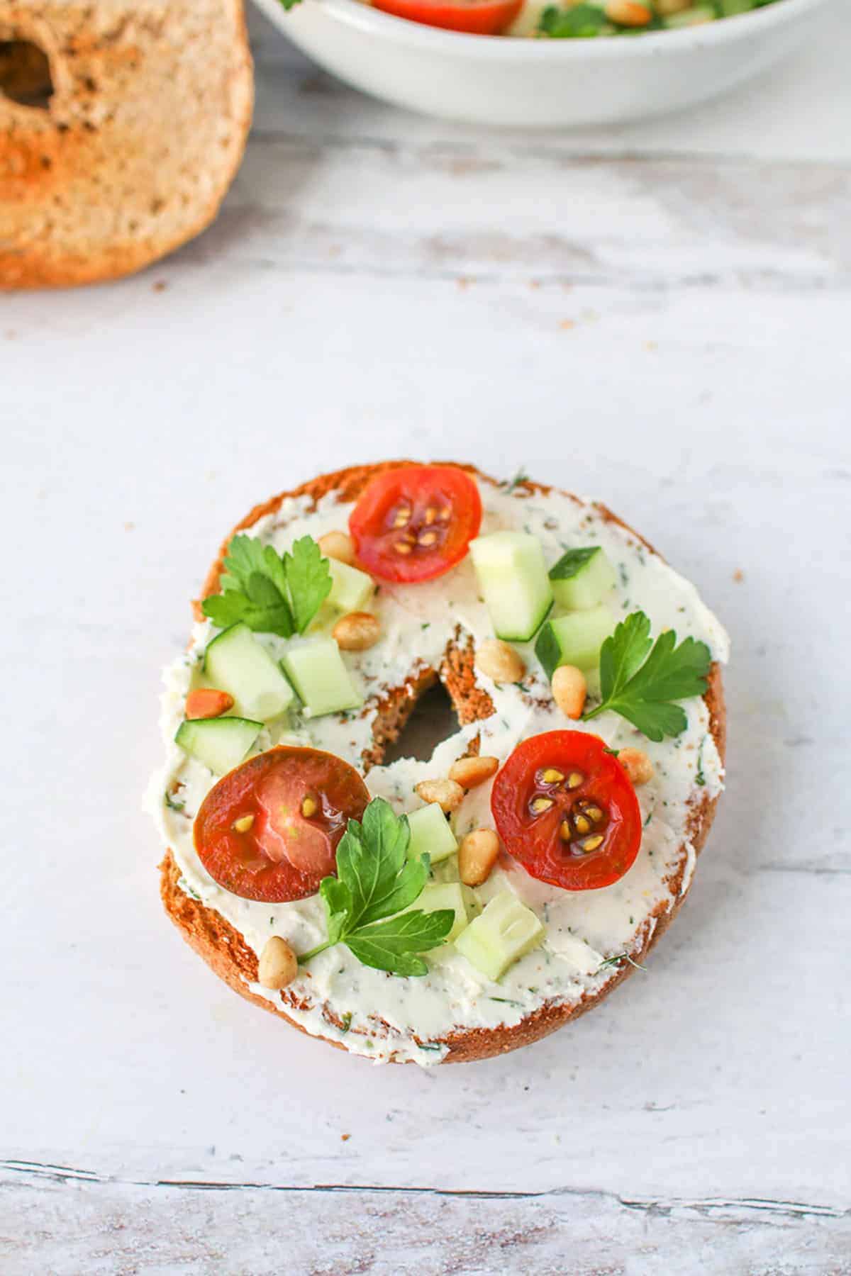A toasted whole wheat bagel with Tzatziki Feta Cream Cheese Spread topped with sliced cherry tomatoes, diced cucumbers, toasted pine nuts and italian parsley leaves