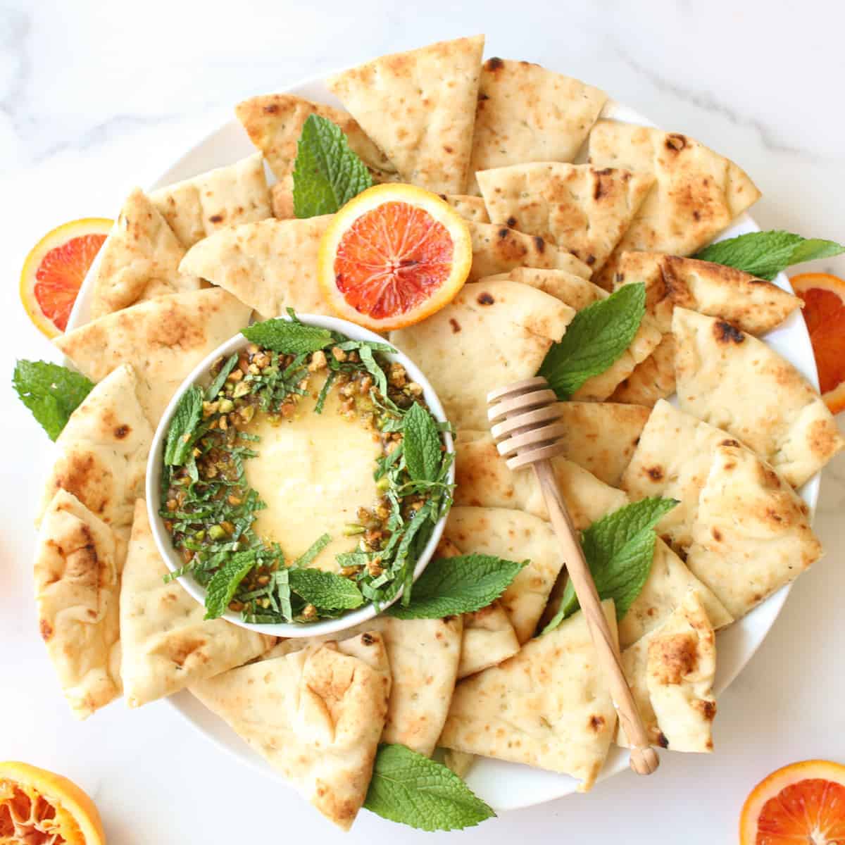 A white platter with triangles of pita and a small bowl with whipped feta, honey, mint and pistachios. Slices of blood orange and mint are around the platter.
