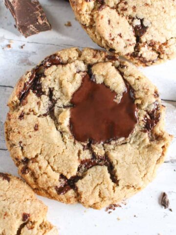 A cookie with a gooey chocolate chunk, the cookies is a light brown on a white and grey background with cookies and chocolate chunks surrounding it.