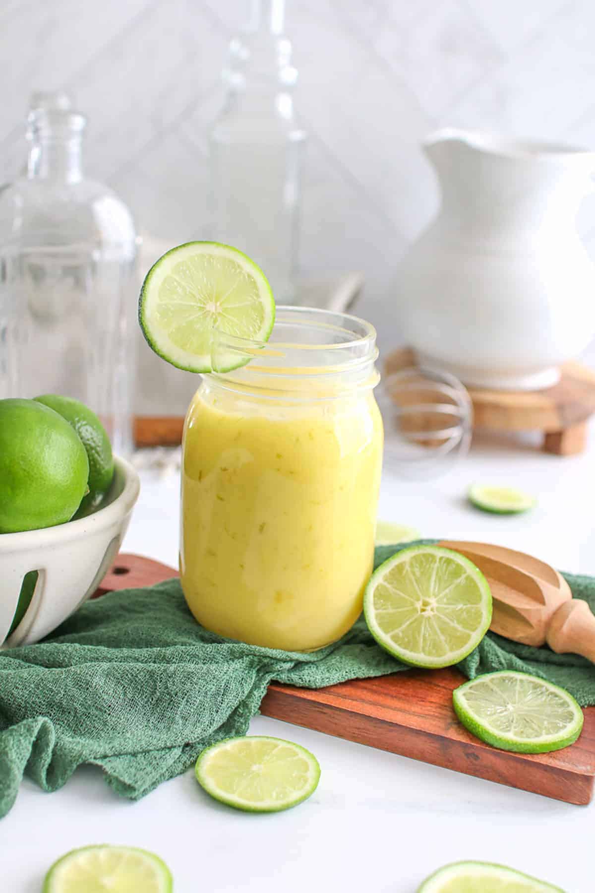 A mason jar fulled of fresh lime curd with a sliced lime on the rim. The jar is on a small wood paddle with limes surrounding it along with a green gauze and citrus zester.