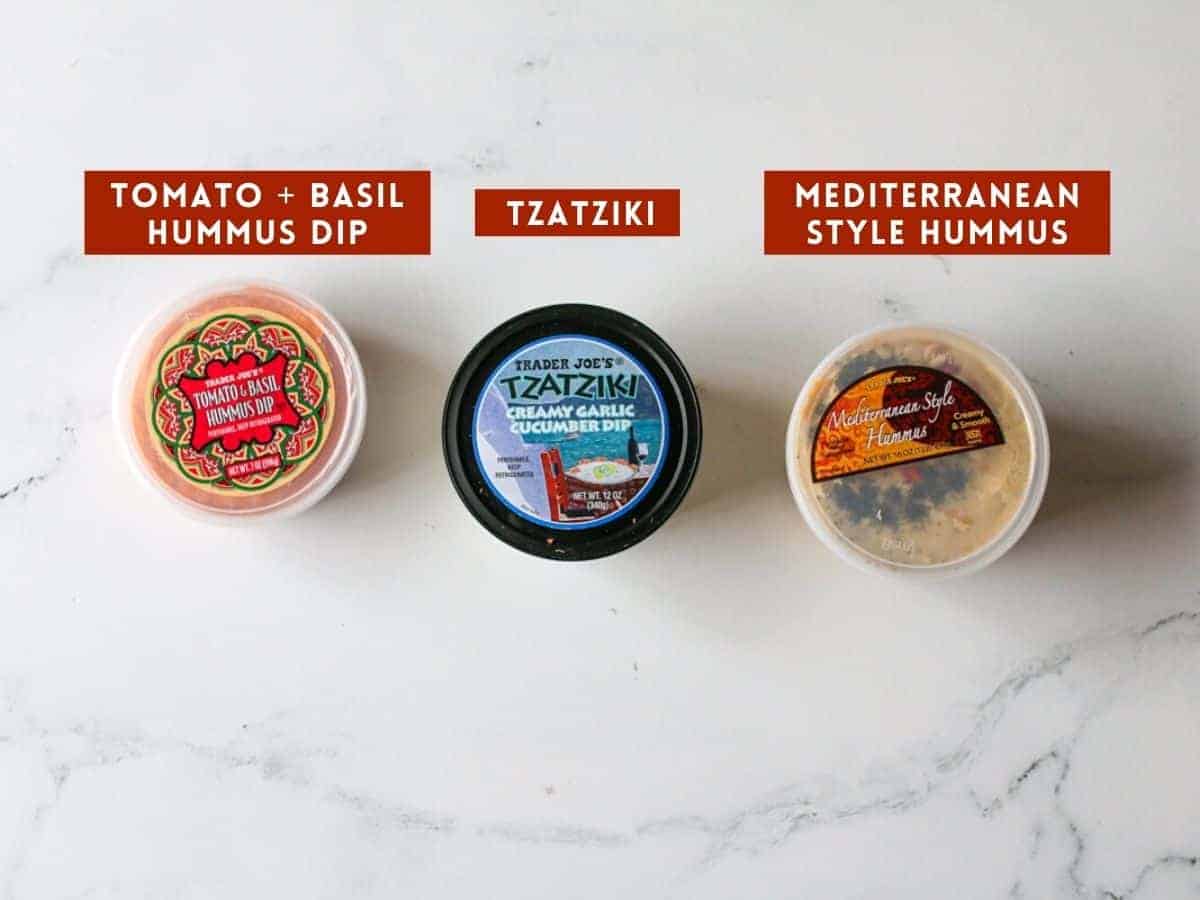 3 different dips in different packages - Tomato & Basil Hummus Dip, Tzatziki Dip and Mediterranean Style Hummus. Each one is labeled with a dark red box with white text capitalized.