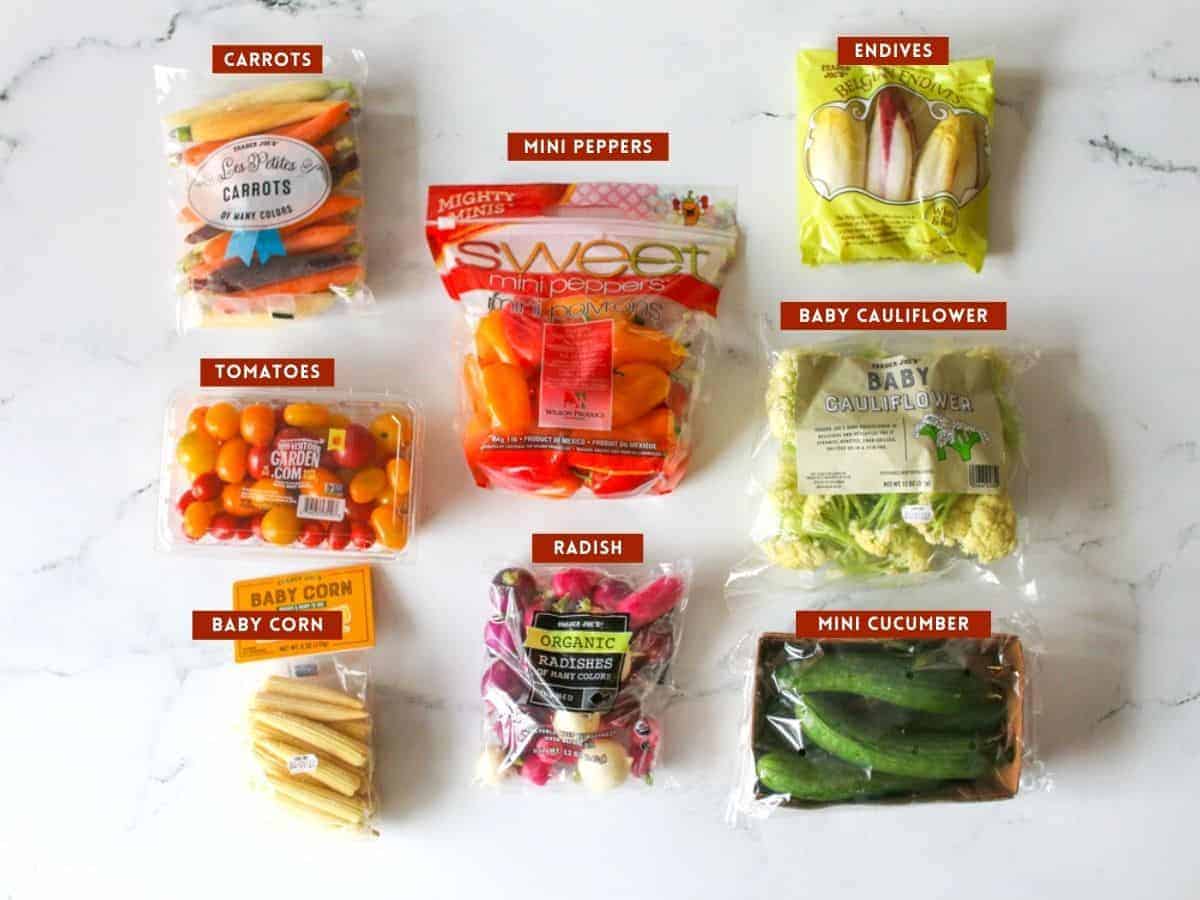 Vegetables in packaging on a white marble background. Each ingredient is labeled with a dark red box with white text capitalized.