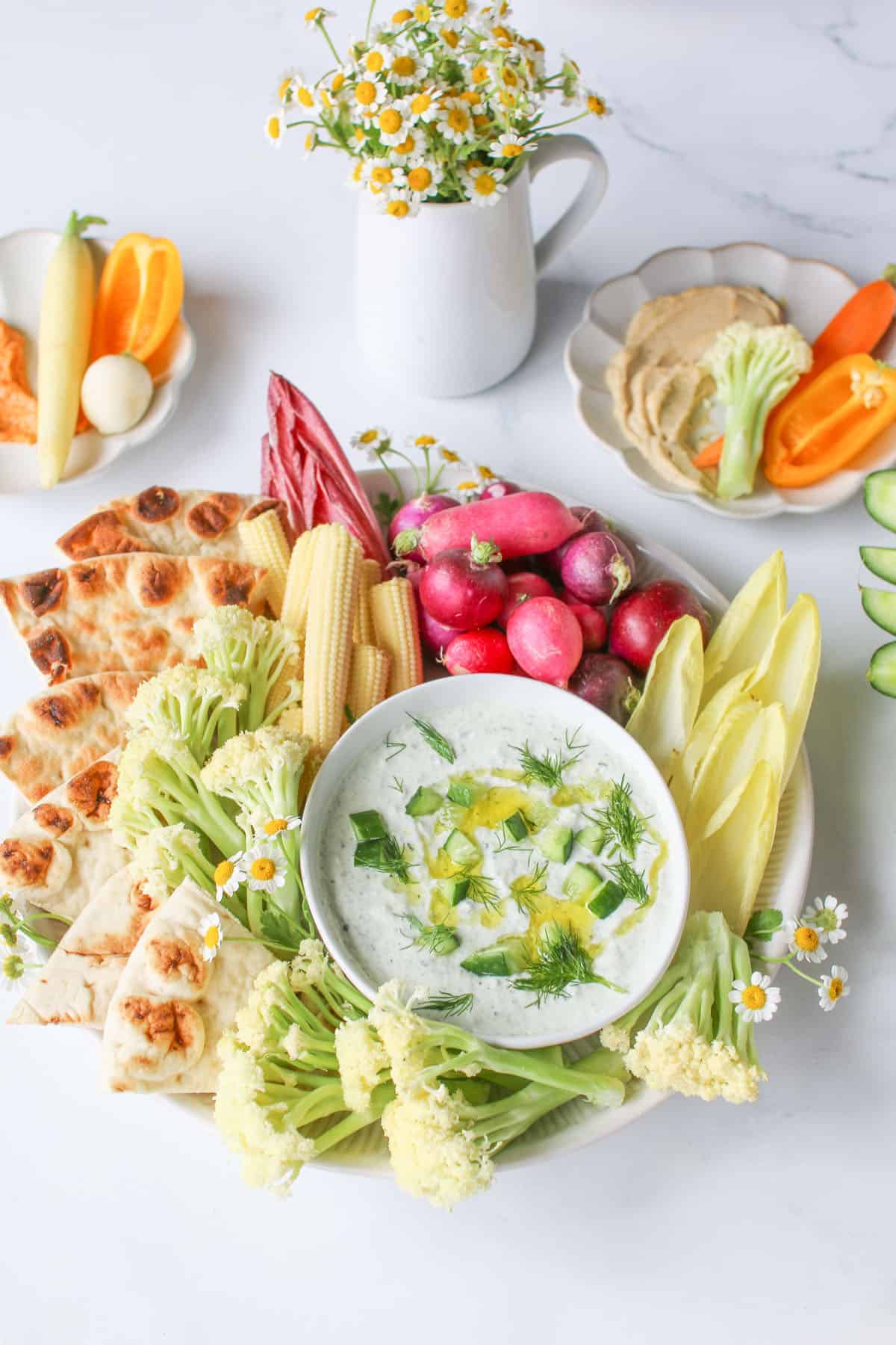 tzatziki dip in a small white bowl with dill, olive oil and diced cucumber on a round plate with vegetables and toasted pita surrounding on a white marble background.