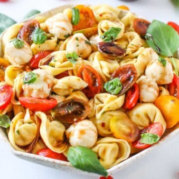 Tortellini Caprese Salad in an octagon bowl with brown speckles with tomatoes, mozzarella and basil leaves on a white marble background with cherry tomatoes surrounding.