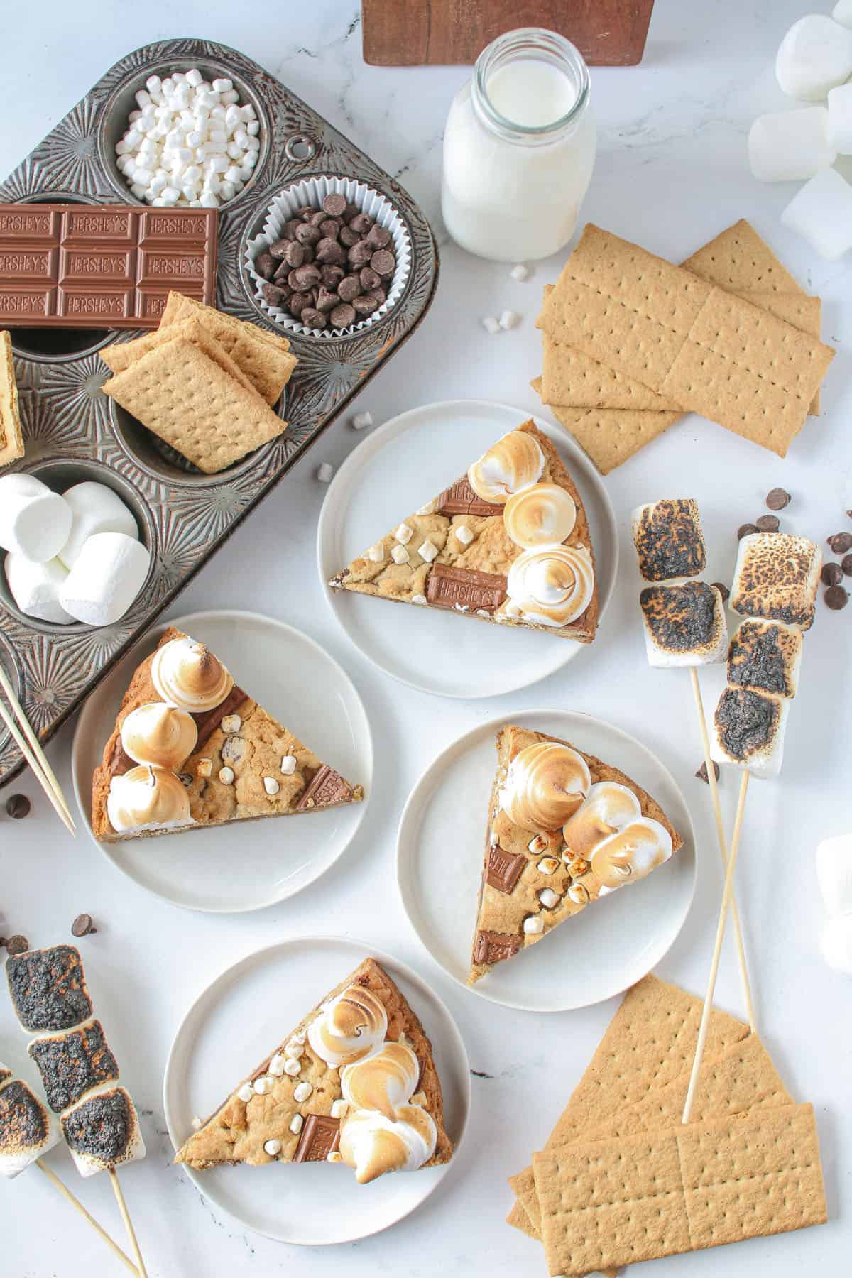 Sliced S'mores Cookie Cake on small white plates on a white marble background with s'mores ingredients in surrounding the ingredients- sheets of graham crackers, chocolate chips, toasted marshmallows on sticks.
