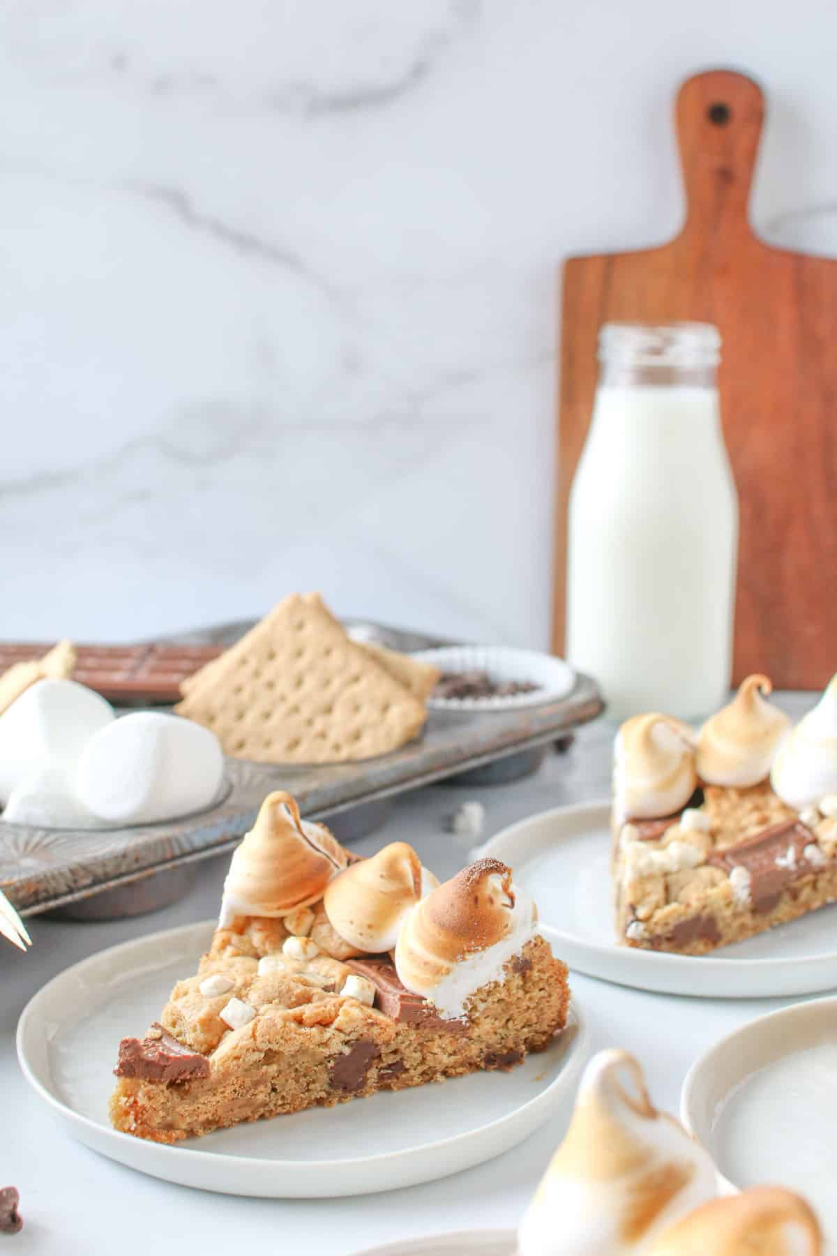 Sliced S'mores Cookie cake on small white circle plates with a muffin tin of s'mores ingredients, a milk bottle of milk and wood paddle in the background.