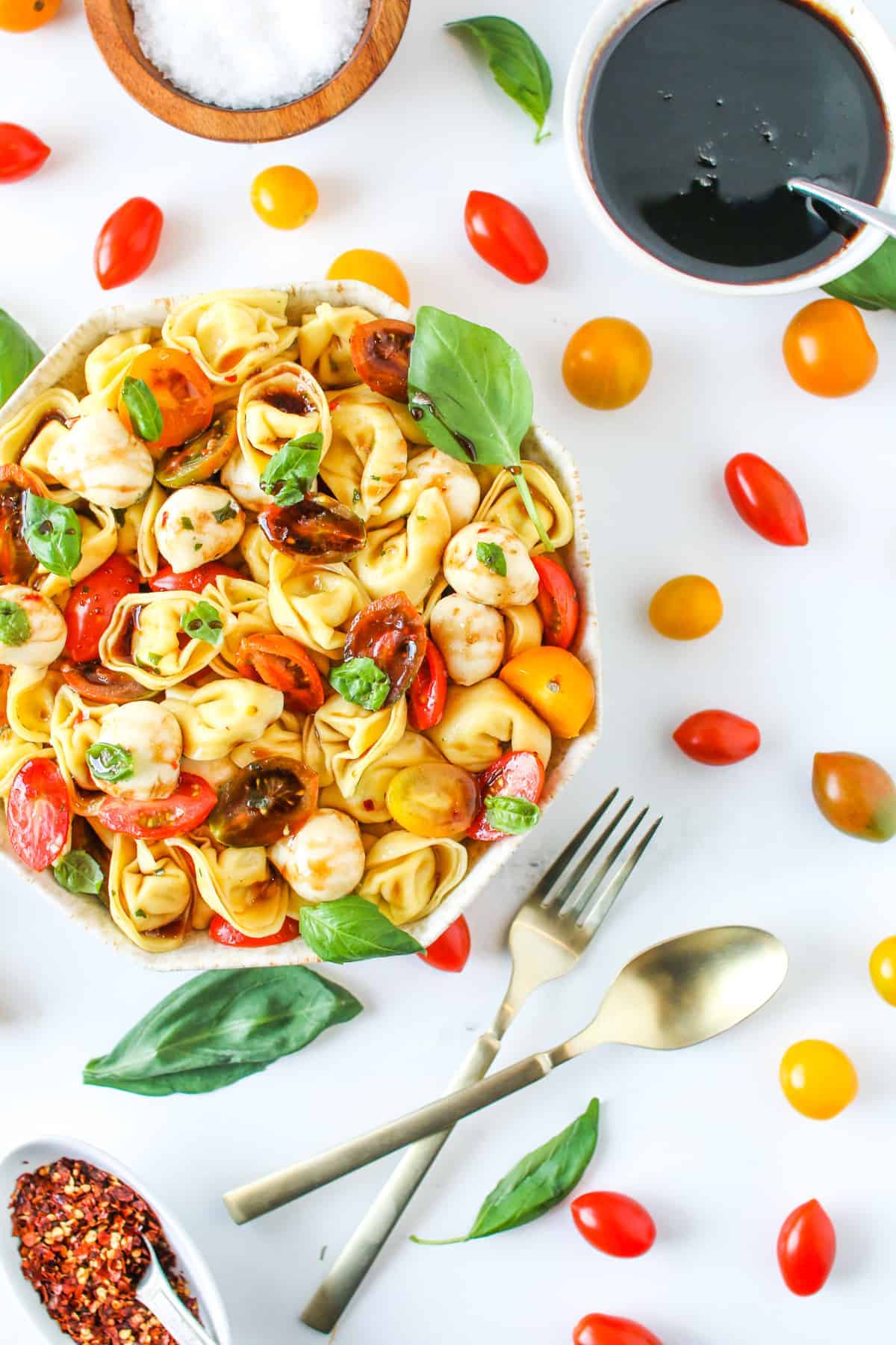 Tortellini Caprese Salad in an octagon bowl with brown speckles with tomatoes, mozzarella and basil leaves on a white marble background with cherry tomatoes, basil, chili flakes in a small white bowl, a gold fork and spoon in the surrounding.
