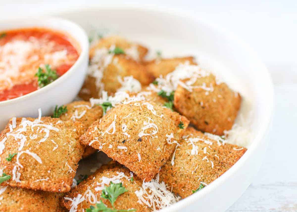 Frozen toasted ravioli in air fryer in a bowl with a small bowl of marinara sauce topped with parmesan cheese and parsley.